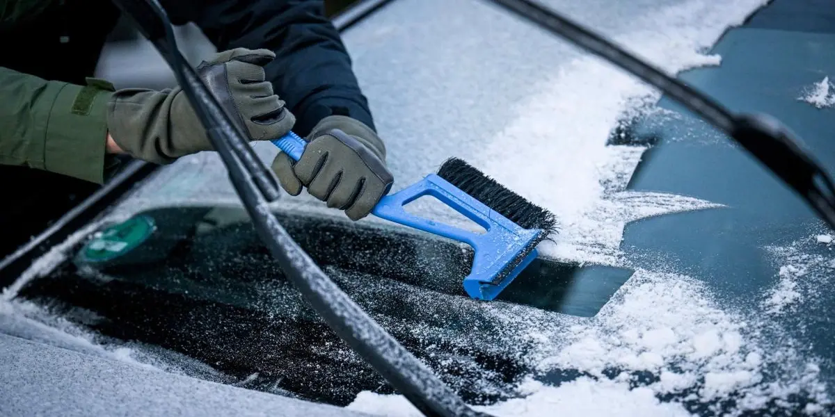 Hand holding a blue ice scraper clearing frost and ice off a frozen windshield.