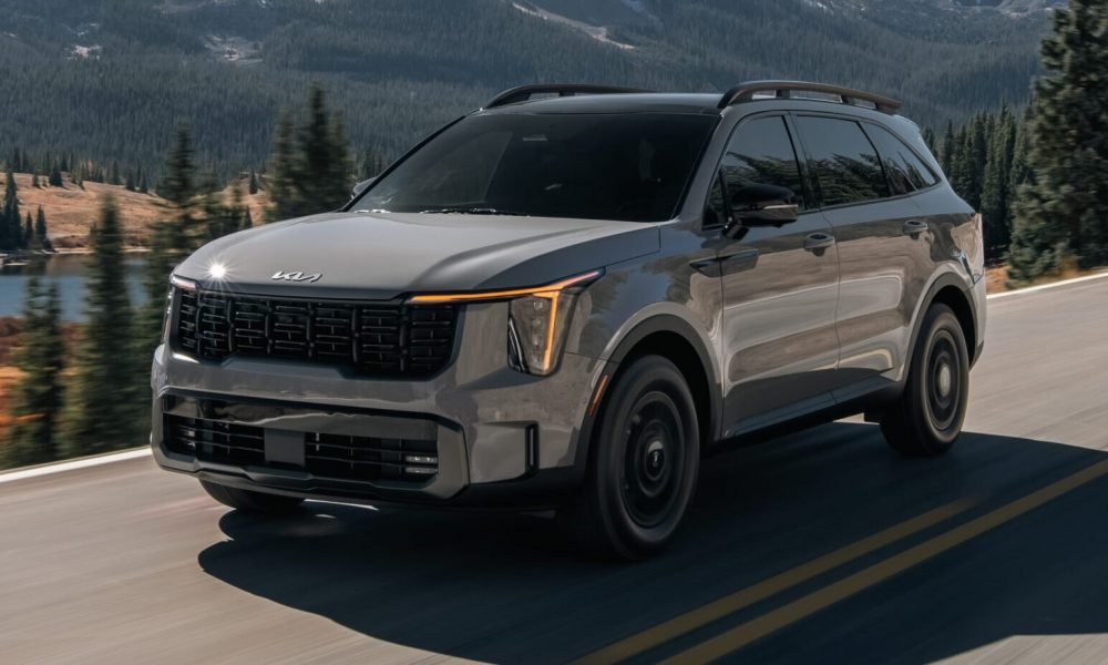 Gray Kia Sorento SUV driving along a mountain road with a forested mountain landscape and lake in the background.