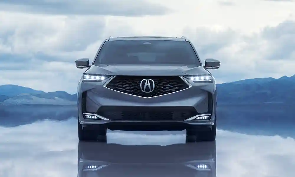 Gray Acura MDX SUV shown from the front, parked on a reflective surface with mountains and a cloudy sky in the background.