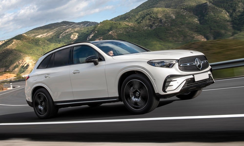 White Mercedes-Benz GLC SUV driving on a road with trees and cloudy skies in the background.