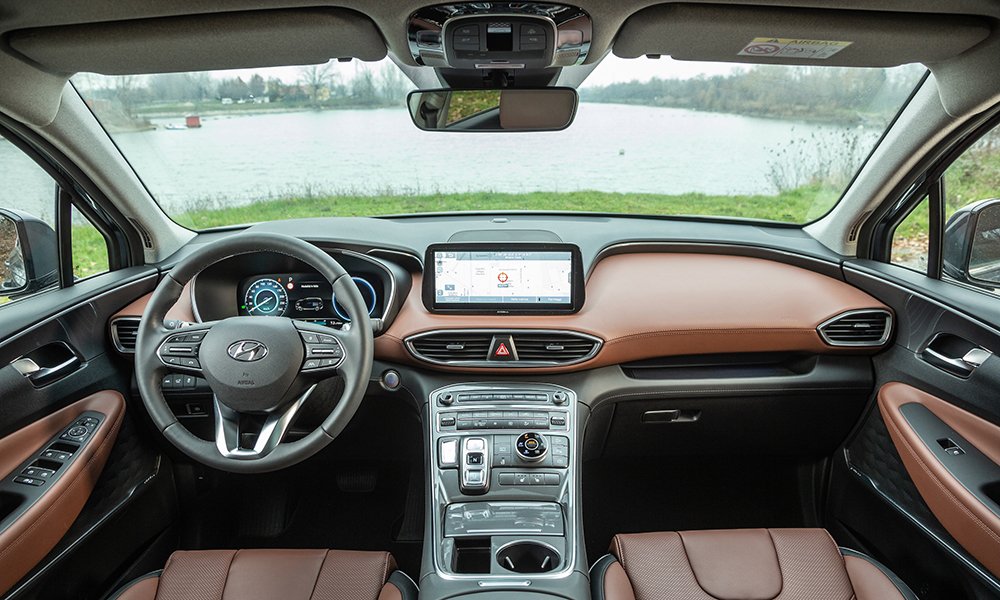 Hyundai Santa Fe interior showing the front dashboard, steering wheel, digital instrument cluster, touchscreen display, and brown leather seats, with a lake visible through the windshield.