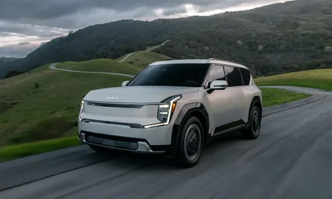 A white Kia EV9 driving along a paved winding road through rolling green hills under an overcast sky.