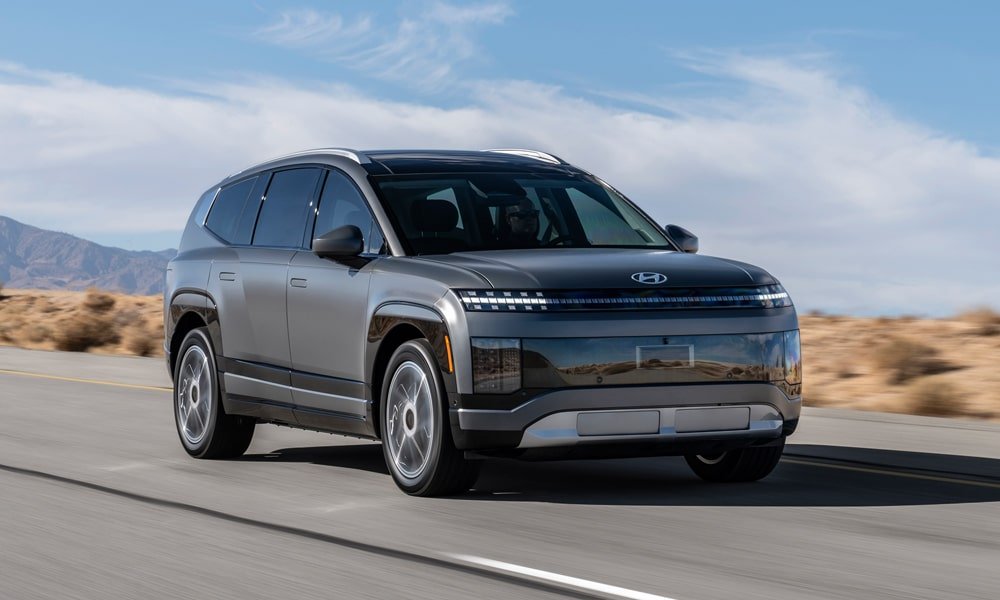 A matte gray Hyundai Ioniq 9 electric SUV driving along a open desert road under a partly cloudy sky.