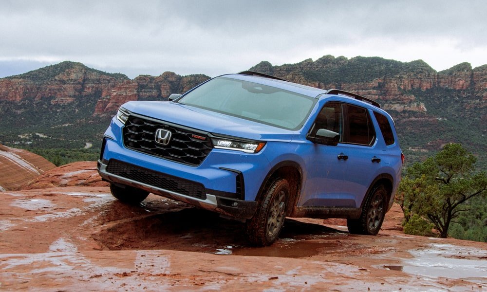 Blue Honda Pilot SUV parked on a rocky trail with red cliffs and mountains in the background.