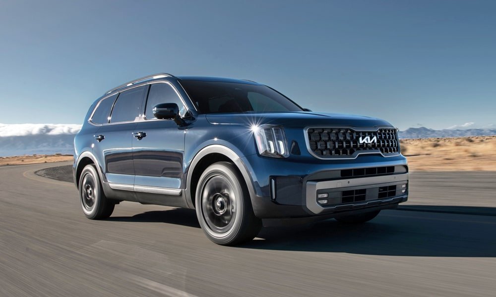 A blue Kia Telluride SUV driving along a paved road through a desert environment.