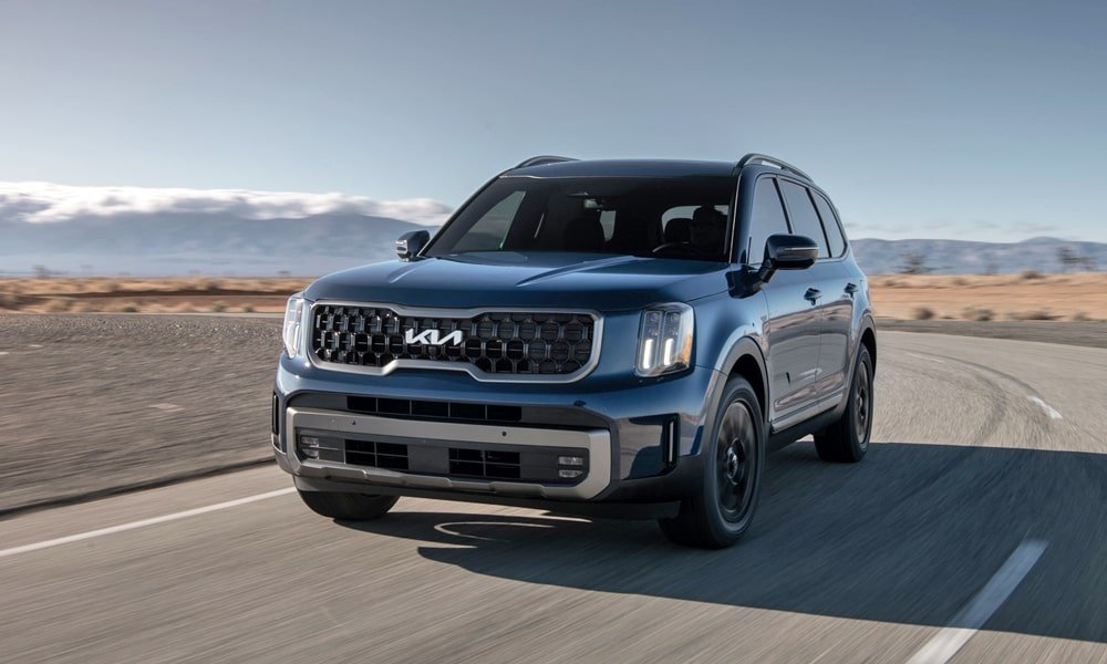 Blue Kia Telluride SUV driving on a paved road through an open desert landscape with mountains in the background.