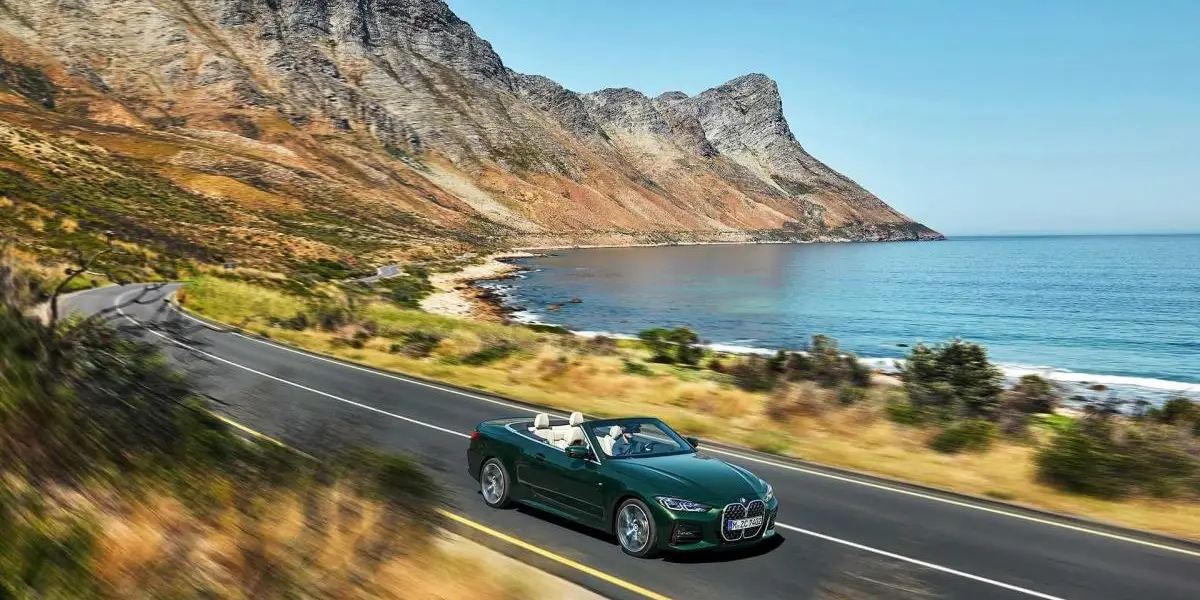 A dark green BMW 4 Series Convertible with an open fabric roof driving along a coastal road next to the ocean