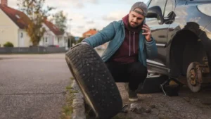Driver preparing to change a flat tire beside a parked car.
